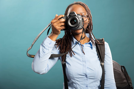 Professional photographer attentively working with her dslr in clean, blue background studio. African american woman wearing backpack, poses with her camera, capturing shots for client.の写真素材