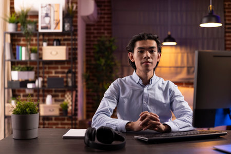 Serious young man sits at desk in home office ready to study online, research on internet and complete freelance tasks. Portrait of asian male entrepreneur with hands clasped, looking at camera.の写真素材