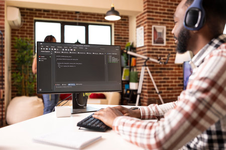 Remote male programmer sits at desk with headphones, coding and debugging scripts on computer. African american man listens to music and works on desktop pc, creating web application from home office.の写真素材
