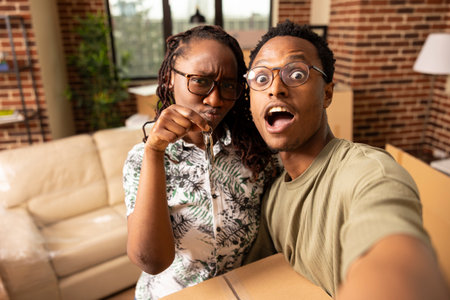 Young african american couple share unpacking humor in new apartment living room. Black woman proudly holds house keys as boyfriend balances box, taking fun selfie together during moving day.の写真素材