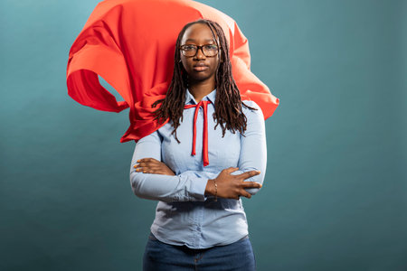African american woman standing tall in power stance, red cape flowing behind. Her focused expression against blue background captures themes of leadership, courage and empowerment.の写真素材