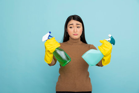 Confused female janitor holding two spray bottles, deciding between detergent solutions. Asian housekeeper wearing yellow gloves, preparing for efficient cleaning service and household chores.の写真素材
