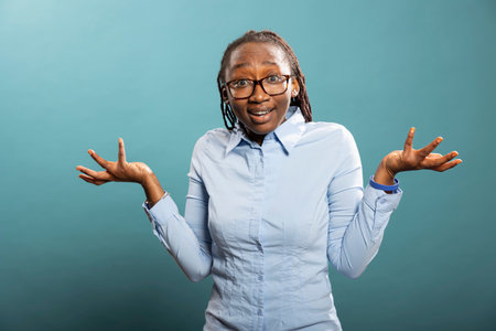 Studio portrait of african american woman with confused expression, raising her shoulders and hands in questioning gesture. Black female model appearing puzzled, standing against blue background.の写真素材