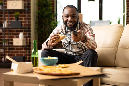 Happy black man holds pizza slice and remote control, increasing TV volume while enjoying good movie. Relaxed weekend moment in modern brick wall apartment, full of comfort and joy.の写真素材