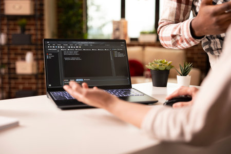 Closeup on laptop displaying programming language as multiethnic people discuss on building an app from home. Diverse couple brainstorming and debugging code for web development in modern apartment.の写真素材