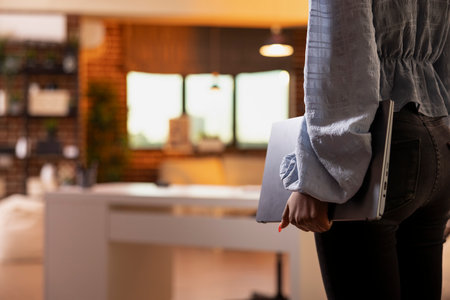 Close up of black woman carrying laptop and walking into room, ready to begin remote work. African American lady holds digital device in home apartment, preparing to handle business tasks after sunsetの写真素材