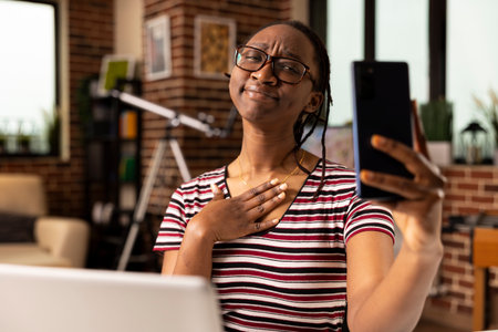 African american female places hand on chest with thankful expression while speaking to colleagues through cellphone. Remote freelancer showing appreciation during virtual meeting.の写真素材