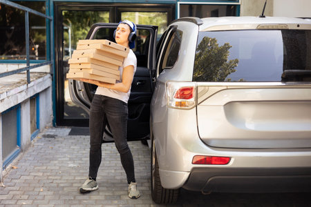 Woman takeout employee delivering boxes of pizza at a building, driving the delivery car for a...