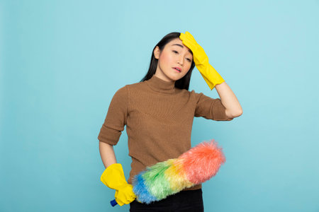 Exhausted asian housekeeper in yellow rubber gloves holding feather duster, showing tired expression...