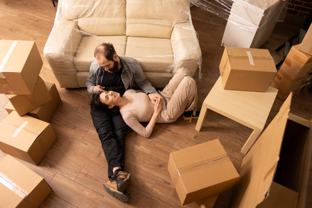 Young couple enjoy bonding on living room floor during moving day. Beautiful woman rests on lap of partner, surrounded by furniture and cardboard boxes, embracing relocation and new home ownership.の写真素材