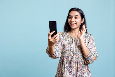 Smiling woman having friendly conversations during teleconference meeting using smartphone, studio background. Indian person having fun catching up with friends during web video call.の写真素材