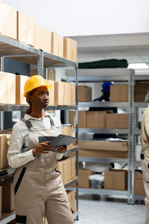 African american girl in safety gear checking merchandise for dispatch in a busy warehouse. Racks and boxes arranged for logistics and transportation in operational supply chain hub.の写真素材