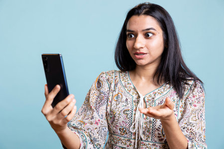 Smiling woman having friendly conversations during teleconference meeting using smartphone, studio background. Indian person having fun catching up with friends during web video call.の写真素材