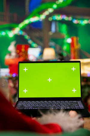 Close up of man in Christmas costume using mockup laptop to browse internet websites, shopping for sleigh accessories. Santa Claus in North Pole workshop searches for sleighs on green screen notebookの写真素材
