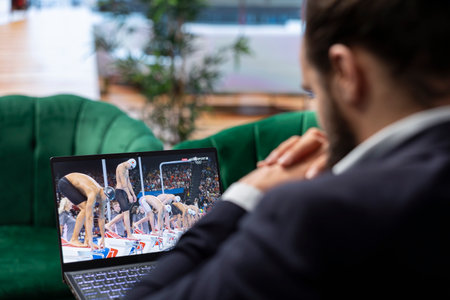 Bucharest, Romania - July 30th 2024 - Young man watching Olympic games online on stream with his laptop, cheering for the national champion at the swimming pool challenge. Olympics live.のeditorial素材