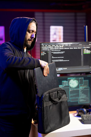 Hacker in hideout putting laptop in backpack after stealing sensitive information using VPN on dark...