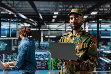 Black military engineer in data center uses laptop for AI systems monitoring, ensuring hybrid warfare operations in server farm army base. Male operator handles machine learning operations.の写真素材