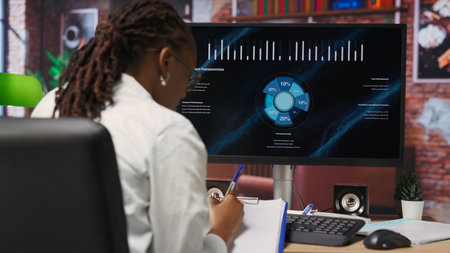 Remote worker at home watching business conference on PC monitor, looking over statistics on documents. Freelancer in apartment looking at presentation and checking paperwork files, camera Bの写真素材