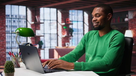 Black teleworker at home desk opening laptop to edit documents and solve daily tasks, updating...