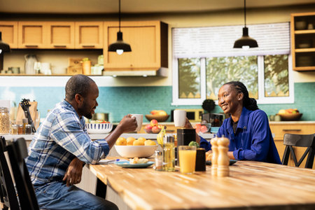 African american lovers clinking mugs with coffee in the morning, enjoying weekend bonding and sitting together. Sweet affectionate man and woman chatting at the breakfast table.の写真素材