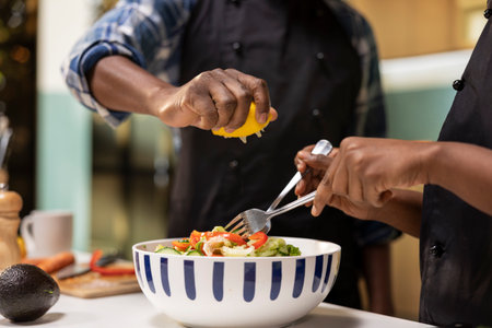 Young black couple adding lemon juice over salad and mixing goods in bowl, enjoying meal prep at...