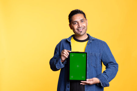 Smiling indian adult presenting digital tablet with isolated mockup screen, staying against studio...