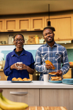 Portrait of black man and woman standing close at the counter kitchen, enjoying fresh coffee and homemade croissants with honey. Joyful couple serving a delicious breakfast with pastry.の写真素材