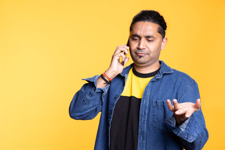 Indian adult chatting with people on remote telephone line in studio, discussing with his friends or close ones against orange background. Young confident joyous man with man bun on a phone call.の写真素材