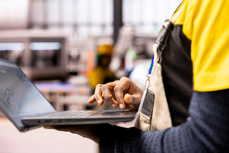 Industrial automation expert ensures efficiency in smart manufacturing plant, using industry 4.0 software on laptop. African american engineer evaluates robotic arms and systems in automated factory.の写真素材