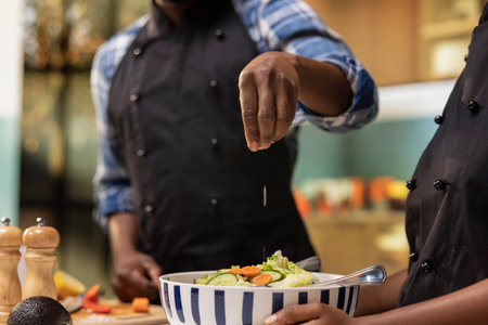 Close up of black people adding pinch of salt to the salad bowl in the kitchen, preparing meal with tasty organic produce. Boyfriend imitating the chef from social media trends, positive energy.の写真素材