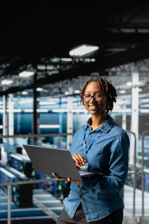 Portrait of black woman engineer solves maintenance tasks on laptop in high tech server hub,...