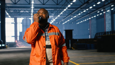 Black operator talks through walkie talkie and monitoring metal fabrication equipment in workshop,...