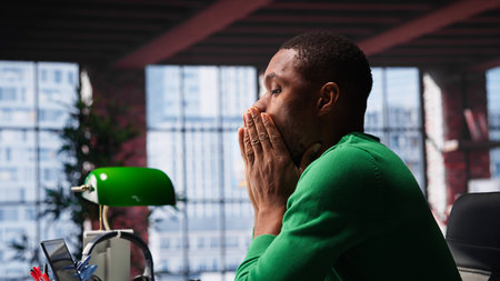 Man feeling exhausted and in pain from lack of sleep at home desk, suffering from insomnia fatigue. Exhausted black guy with negative emotion affected by burnout, feeling stressed.の写真素材