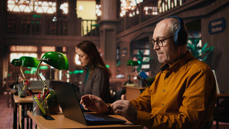Elderly person does dance moves with songs on headset in the library, learning for academic coursework and listening to music. Senior man singing and dancing next to green lamp.の写真素材
