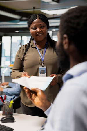 Smiling employee hands over documents to colleague in modern office setting. Happy african american woman exchanging paperwork with coworker, concept of friendly workplace interactionの写真素材