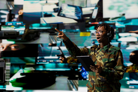 Shocked army dispatcher soldier in camouflage gasps while pointing to screen detecting enemy activity. Frantic military dispatcher wearing headset reacts to enemy threat on digital displayの写真素材