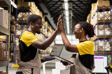 Team of pleased workers sharing a high five gesture in industrial depot with huge storage racks, proud of teamwork success for the distribution operations. Efficiency achievement.の写真素材