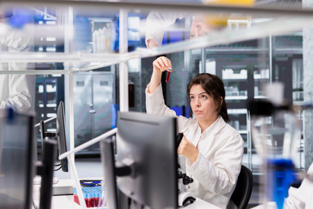 Woman looking at blood sample in test tubes in high tech laboratory workspace. Clinical lab researcher analyzing sanguine fluid specimen in vial used for medical researchの写真素材