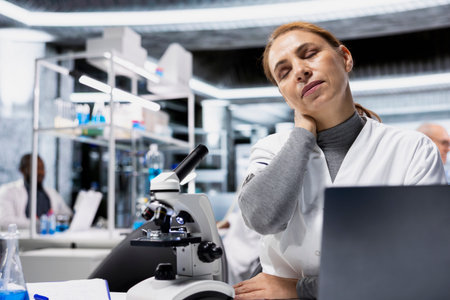 Exhausted scientist in lab suffering neck ache while documenting clinical trial outcomes on laptop....