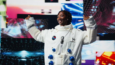 Happy astronaut in futuristic space suit doing dance moves against vibrant cosmic visuals backdrop. Playful african american woman dancing, having fun during space exploration, camera Aの写真素材