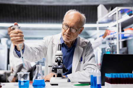 Senior biochemist in lab adding drops of liquid into blue solution to refine new synthetic materials. Elderly man in laboratory mixing chemicals in test tube to watch for reactionの写真素材