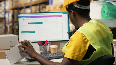 Black depot clerk reviewing inventory data on tracking software for distribution system, busy...