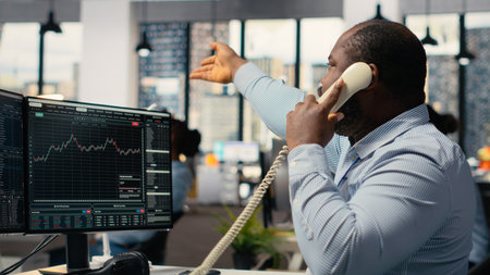 Distressed brokerage firm trader making phone calls to clients in response to Black Swan event shock. African american man evaluate stock exchange impact of unexpected recessionの写真素材