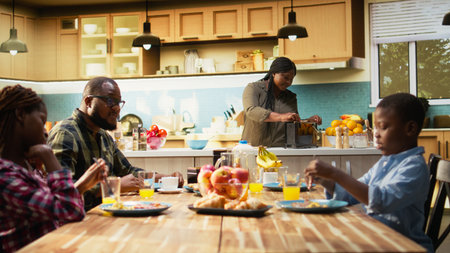 African American mother preparing breakfast in the morning light while the family wait at the table with croissants, fruits and orange juice. Parents and children laugh together, love. Camera A.の写真素材