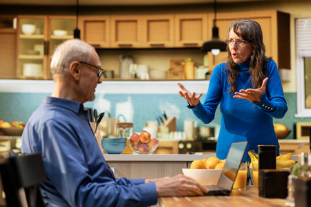 Senior couple experiences a heated quarrel in the kitchen, woman enraged and man upset by the disagreement. Aggressive shouting and confrontation reflect lifestyle problems during retired life.の写真素材