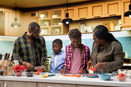 Joyful African American parents teaching kids to cook and cut vegetables safely, family enjoys...