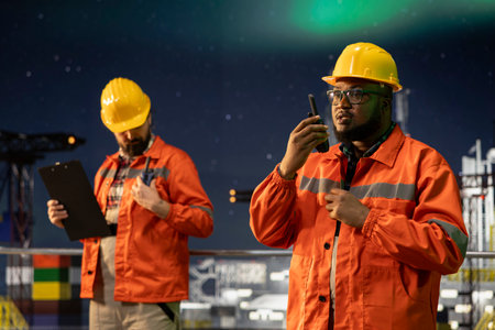 Radio operator working on drilling derrick on ocean provides operational updates with walkie talkie. African american man on offshore platform maintains radio contact at night under aurora borealisの写真素材