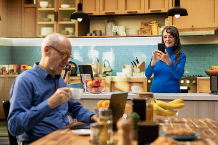 Senior adult browsing internet on smartphone and smiles in kitchen, relaxing with coffee and...