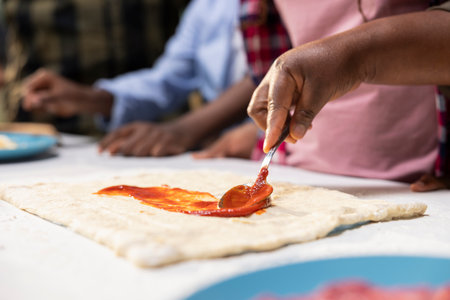 Close up of children spreading tomato sauce on pizza dough at home, making homemade meal with fresh ingredients and produce. Adding dressing as first step, family time in the kitchen.の写真素材