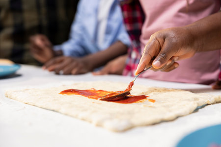 Close up of african american parents and kids using tomato sauce on the dough, preparing a homemade...